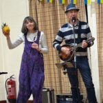 Man in black and grey stripey shirt and woman in purple dungarees playing musical instruments